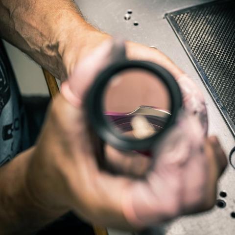 Photographie montrant la main d'un ouvrier tenant une loupe à travers laquelle on peut apercevoir un boîtier de montre.